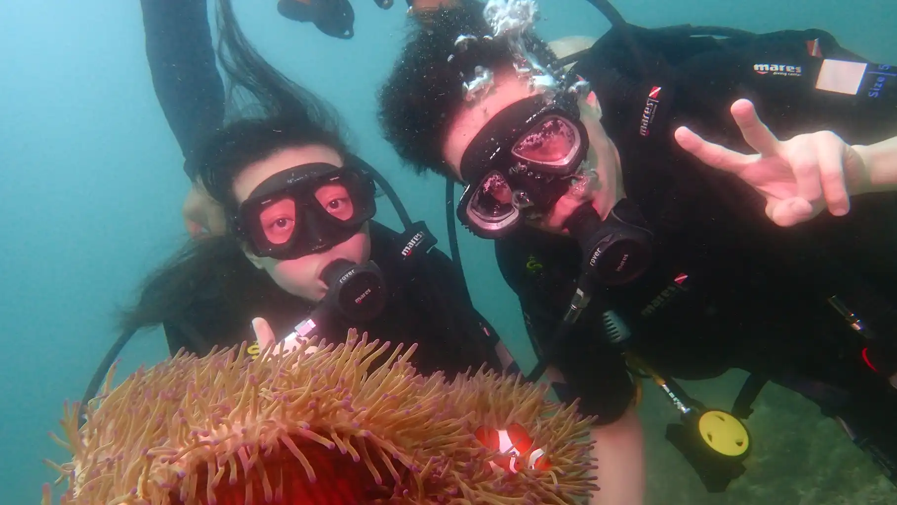 Two scuba divers underwater with a sea anemone and clownfish, making peace signs.
