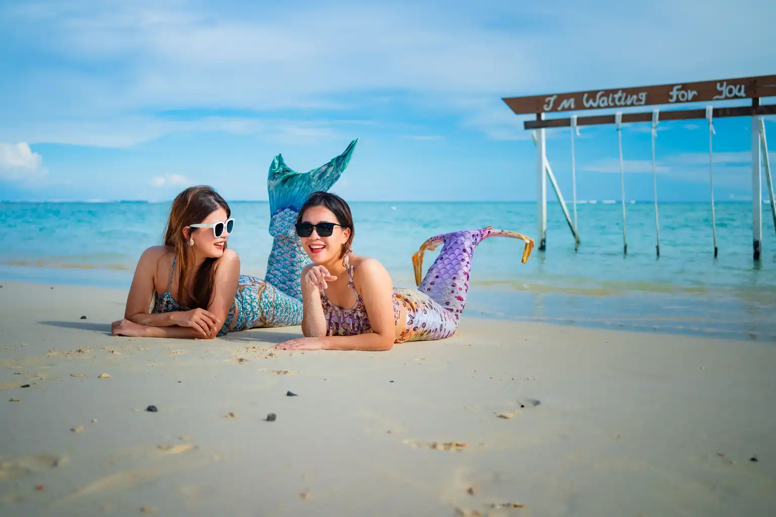 Two women wearing mermaid tails and sunglasses lying on a beach, with a sign saying I'm Waiting for You in the background.